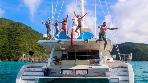A family leaping off the flybridge of a sailing catamaran anchored in a calm bay below a green hillside in the British Virgin Islands