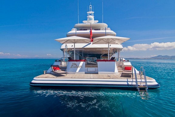 The shaded aft deck and swim platform of the superyacht MY EDEN, with two parasols, red-cushioned loungers and a boarding ladder down into flat blue water