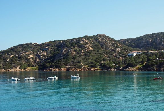 The bay at Baja Sardinia in northern Sardinia seen from the water, with RIBs and tenders on moorings in clear turquoise shallows and a granite headland of scrub and rock rising behind, a stone fort on the skyline