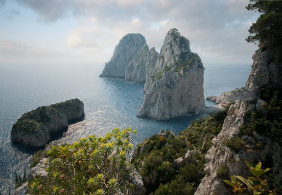 The three Faraglioni limestone sea stacks rising out of the water off the south coast of Capri, seen from the cliffs above, with the arch through the middle stack and open sea beyond