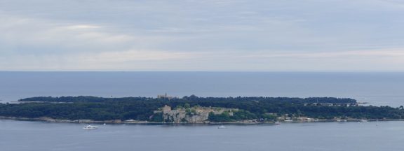 Ile Sainte-Marguerite seen from across the water off Cannes, its low wooded ridge topped by the stone Fort Royal, with Ile Saint-Honorat behind it and yachts anchored in the channel between the island and the mainland