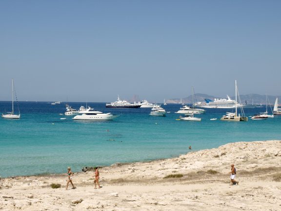 Motor yachts, sailing yachts and a catamaran anchored in the turquoise shallows off Playa de ses Illetes on Formentera, with people walking the low sandy spit in the foreground and the coast of Ibiza on the horizon