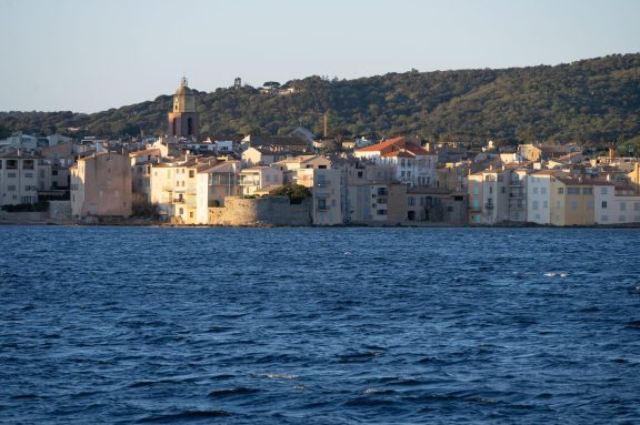 Saint-Tropez seen from the water in low evening light, the ochre and pink houses of the old town lining the shore beneath the bell tower of Notre-Dame de lAssomption, with wooded hills rising behind