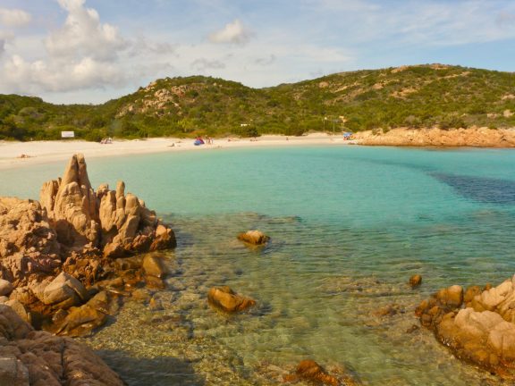 Spiaggia del Principe on Sardinias Costa Smeralda, with weathered pink granite rocks in the foreground, clear turquoise shallows over a sandy bottom and a curve of pale beach backed by low green maquis